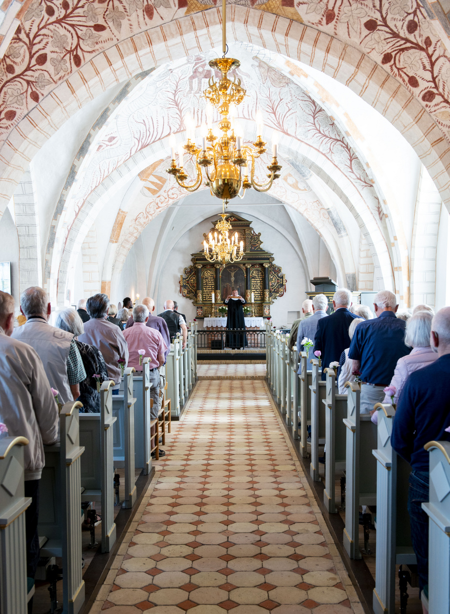Menighed siddende i bænke mod præst og bemalede kirkehvælvinger.