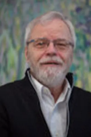 Portræt af en mand med gråt hår, skæg, briller og mørk jakke.