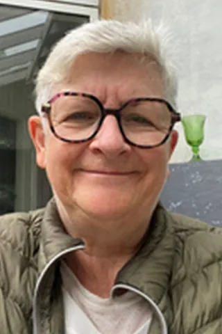 Portræt af en smilende kvinde med kort hvidt hår, briller og grøn jakke.